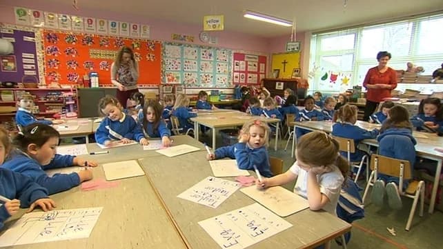 A primary classroom where children in school uniform sit at grouped tables writing their names on cards, with two adults standing at the sides of the room