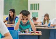 A boy sitting at a classroom desk with his head in his hands while other children work behind him