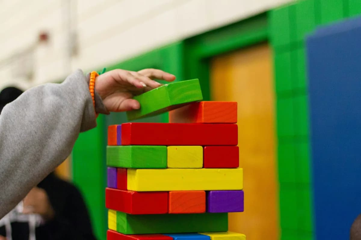 A child's hand carefully placing a green block on a tower of brightly coloured wooden blocks