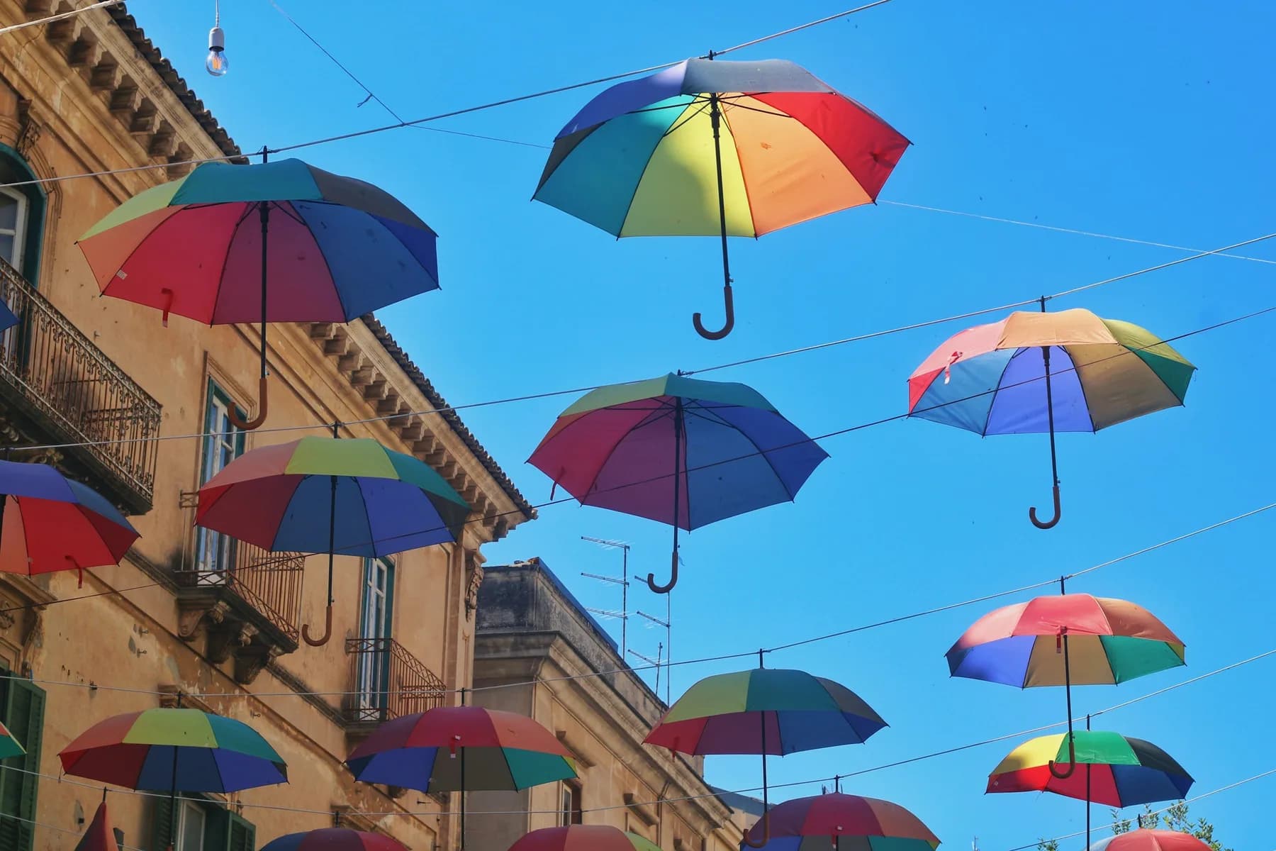 Dozens of rainbow-coloured umbrellas strung on lines above a street, open against a bright blue sky
